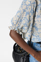 A person wearing a blue and white blouse fluide with a delicate motif floral design and blue jeans holds a black leather bag over their shoulder. The image focuses on their arm, blouse sleeve, and the bag.