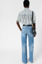 Person facing away, wearing a short-sleeve blouse fluide à motif floral, high-waisted light blue jeans, black belt, and carrying a black shoulder bag against a plain white background.