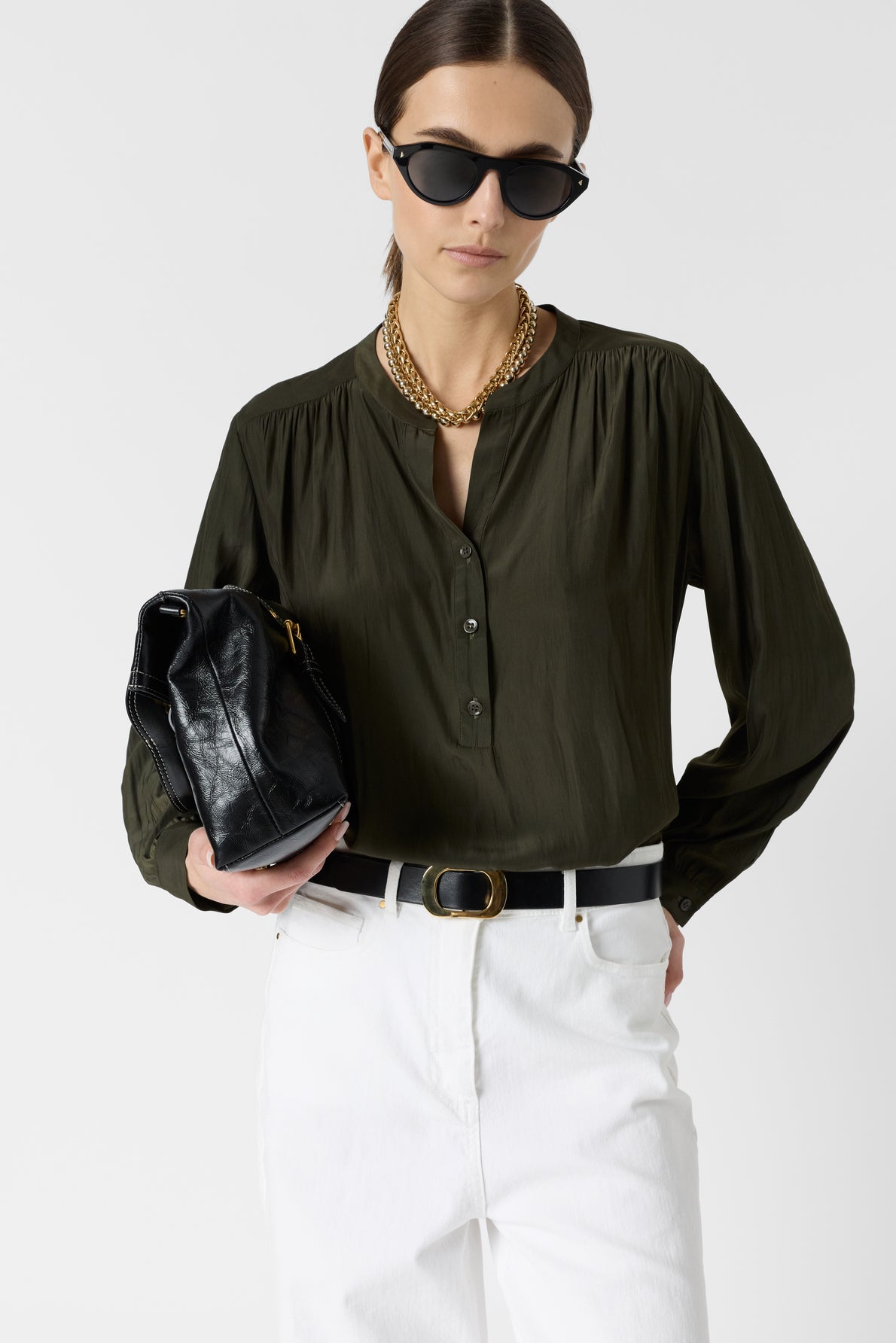 Woman wearing dark sunglasses, a gold necklace, olive green blouse, and white pants, holding a black handbag against a plain background.