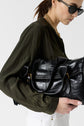 Woman in sunglasses and a gold chain necklace holds a shiny black leather handbag under her arm. She wears an olive green blouse and white pants, set against a plain white background.