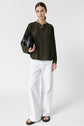 Woman stands against a white background, wearing a dark green long-sleeve blouse, white wide-leg pants, and black sandals. She holds a black clutch and has straight brown hair pulled back.