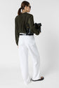 A woman with a brown ponytail, wearing sunglasses, an olive green blouse, white wide-leg pants, and black sandals, holds a black bag while facing away from the camera against a plain white background.