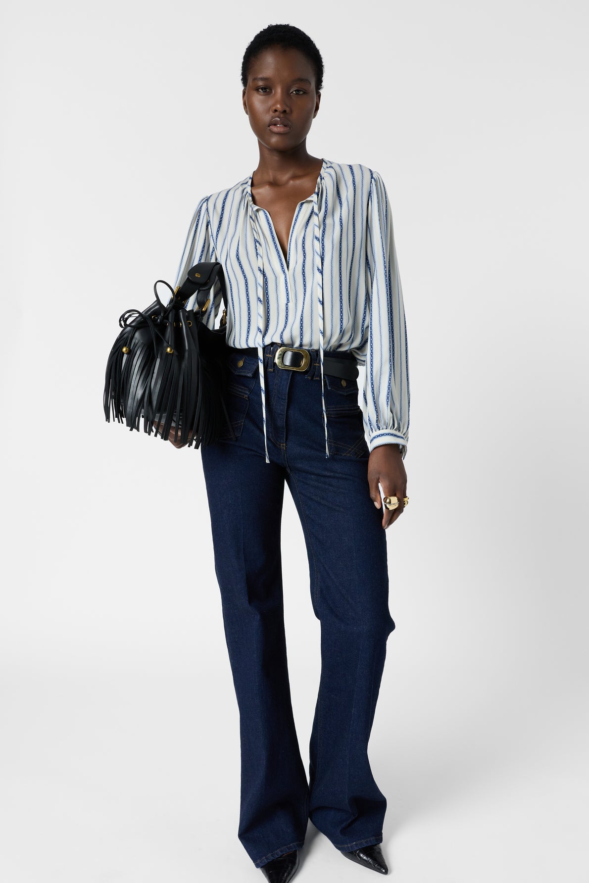 A woman wearing a blouse à rayures, dark flared jeans, and pointed shoes holds a black fringed handbag. She has short hair and stands against a plain white background.