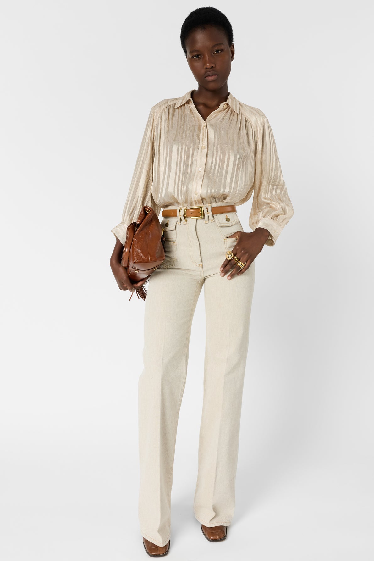 A person stands facing forward, wearing a shiny beige blouse, high-waisted cream pants, and brown shoes. They hold a brown clutch bag and have short hair, accessorized with rings and a belt.