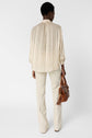 A person stands facing away, wearing a light beige, long-sleeve, pleated top and cream-colored pants, holding a brown leather handbag with tassel details in their left hand. The background is plain white.
