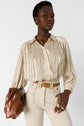 A person wearing a beige striped blouse and high-waisted light pants stands holding a brown leather handbag, accessorized with gold rings and a tan belt, against a plain white background.