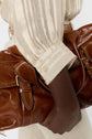 A person with dark skin wearing a beige striped blouse holds a large brown leather handbag with gold buckles close to their body.