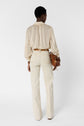 Person standing with back to camera, wearing light beige high-waisted pants, a cream-colored long-sleeve blouse, and holding a brown handbag. Background is plain white.