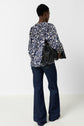 Person facing away, wearing a blue and white patterned top with long sleeves and dark flared jeans, holding a large black fringed handbag. The background is plain and white.