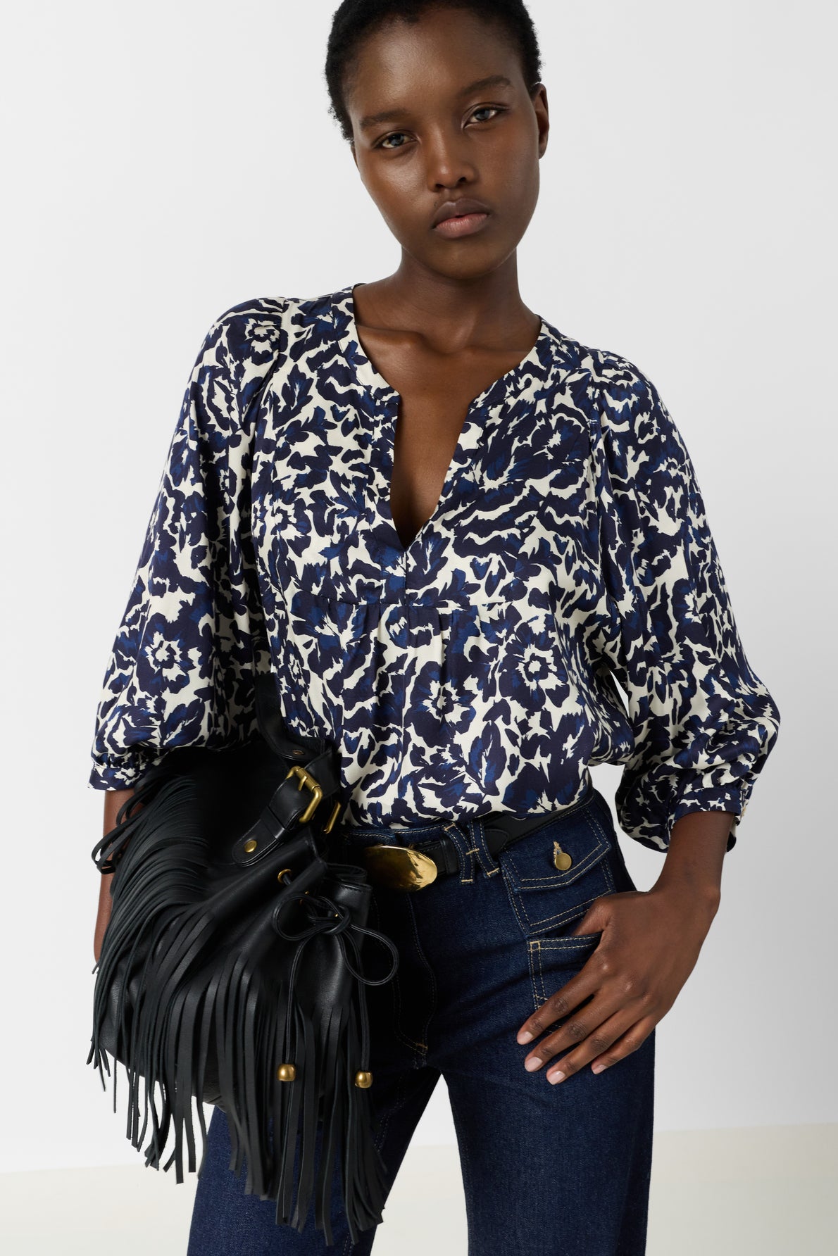 A person wearing a blue and white patterned blouse, dark jeans, and holding a black fringed handbag poses with one hand on their hip against a plain background.