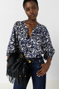 A person wearing a blue and white patterned blouse, dark jeans, and holding a black fringed handbag poses with one hand on their hip against a plain background.