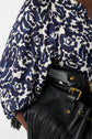 A close-up of a person wearing a blue and white floral-patterned blouse with puffed sleeves, holding a black leather bag with gold hardware and fringe detailing.