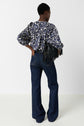 Person standing with back facing the camera, wearing a blue and white patterned blouse, dark blue flared jeans, and holding a black fringed bag over one shoulder against a plain white background.