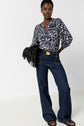 A woman poses in wide-leg dark jeans, a blue and white patterned blouse, black sandals, and a belt with a gold buckle, holding a black fringe bag against a plain white background.