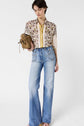 Woman wearing a patterned blouse with yellow accents, light blue high-waisted wide-leg jeans, holding a brown bag, standing against a plain white background.