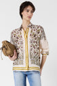 Woman with short brown hair wears a patterned button-up shirt with gold and purple details, light blue jeans, and holds a tan suede bag against a white background.