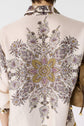Person wearing a light-colored shirt with an intricate, symmetrical floral and paisley pattern in purple, yellow, and brown tones. The image is focused on the back of the shirt and part of a beige handbag is visible.