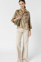 A woman wearing a patterned blouse, beige wide-leg pants, and holding an olive green purse stands against a plain white background.