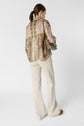 Woman with a ponytail wearing a colorful patterned blouse, cream wide-leg pants, and brown shoes, holding an olive green clutch, standing against a plain white background, facing away from the camera.