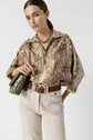 Woman in patterned blouse, cream jeans, and brown woven belt; holding an olive quilted purse, wearing a chunky gold necklace, with hair pulled back, standing against a plain white background.