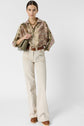 Woman wearing a patterned blouse, cream wide-leg pants, brown belt, brown shoes, gold necklace, and holding a green quilted bag, standing against a plain white background.
