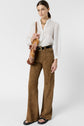 Woman wearing a white chemise brodée with a delicately embroidered col à volants, brown flared pants, black sandals, and a brown shoulder bag, standing against a plain white background.