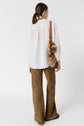 A woman with brown hair in a low ponytail wears a white chemise brodée, brown wide-leg pants, and black shoes, carrying a multicolored woven shoulder bag. She is standing facing away against a plain white background.