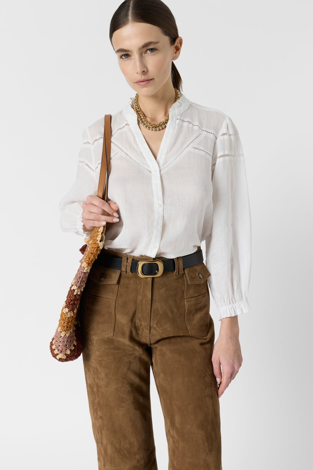 A woman wearing a white blouse with a col à volants, brown suede pants, a chunky gold necklace, and a brown belt stands holding a brown sequin bag over her shoulder against a plain white background.