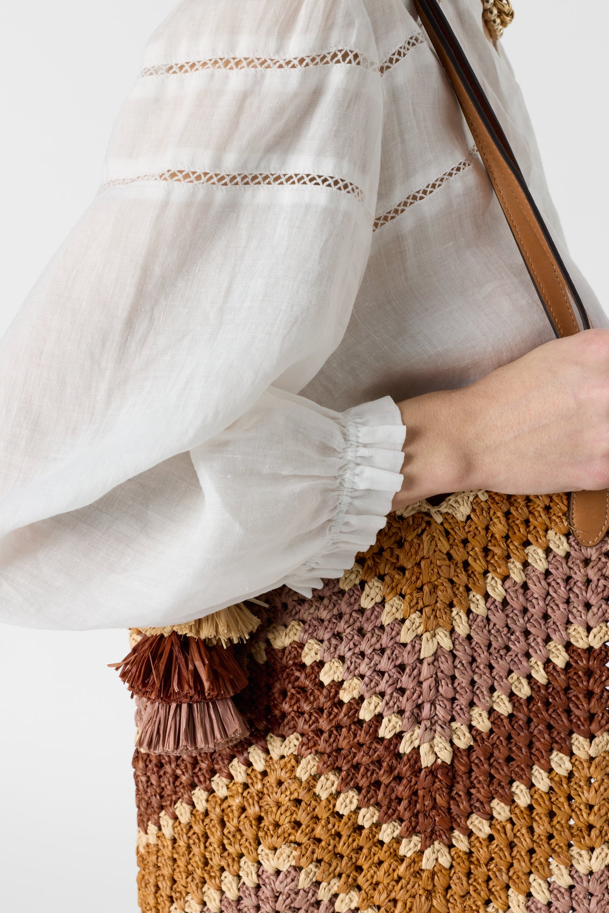 A person wearing a white long-sleeve blouse with col à volants holds a woven, zigzag-patterned straw bag with brown, beige, and tan colors and a tassel detail.