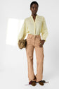 A person wearing a light yellow chemise brodée crafted from tissu fin ramie, tan high-waisted pants with textured side seams, and brown shoes stands against a plain white background, holding a tan handbag.