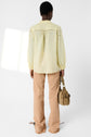 A person stands facing away, wearing a pale yellow long-sleeve blouse crafted from tissu fin and light brown pants, holding a tan handbag in their left hand. The background is plain white.