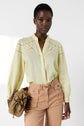 A woman wearing a light yellow chemise brodée in tissu fin and tan high-waisted pants holds a brown bag. She stands against a plain white background, looking confidently at the camera.