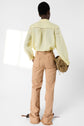 A person stands facing away, wearing a light yellow chemise brodée and tan pants with frayed edges, holding a brown bag. The delicate tissu fin of the blouse catches the light streaming in from the left, set against a plain white background.