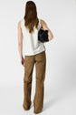 A person with long brown hair, wearing a sleeveless white top and brown pants, stands facing away while holding a black bag under their left arm against a plain white background.
