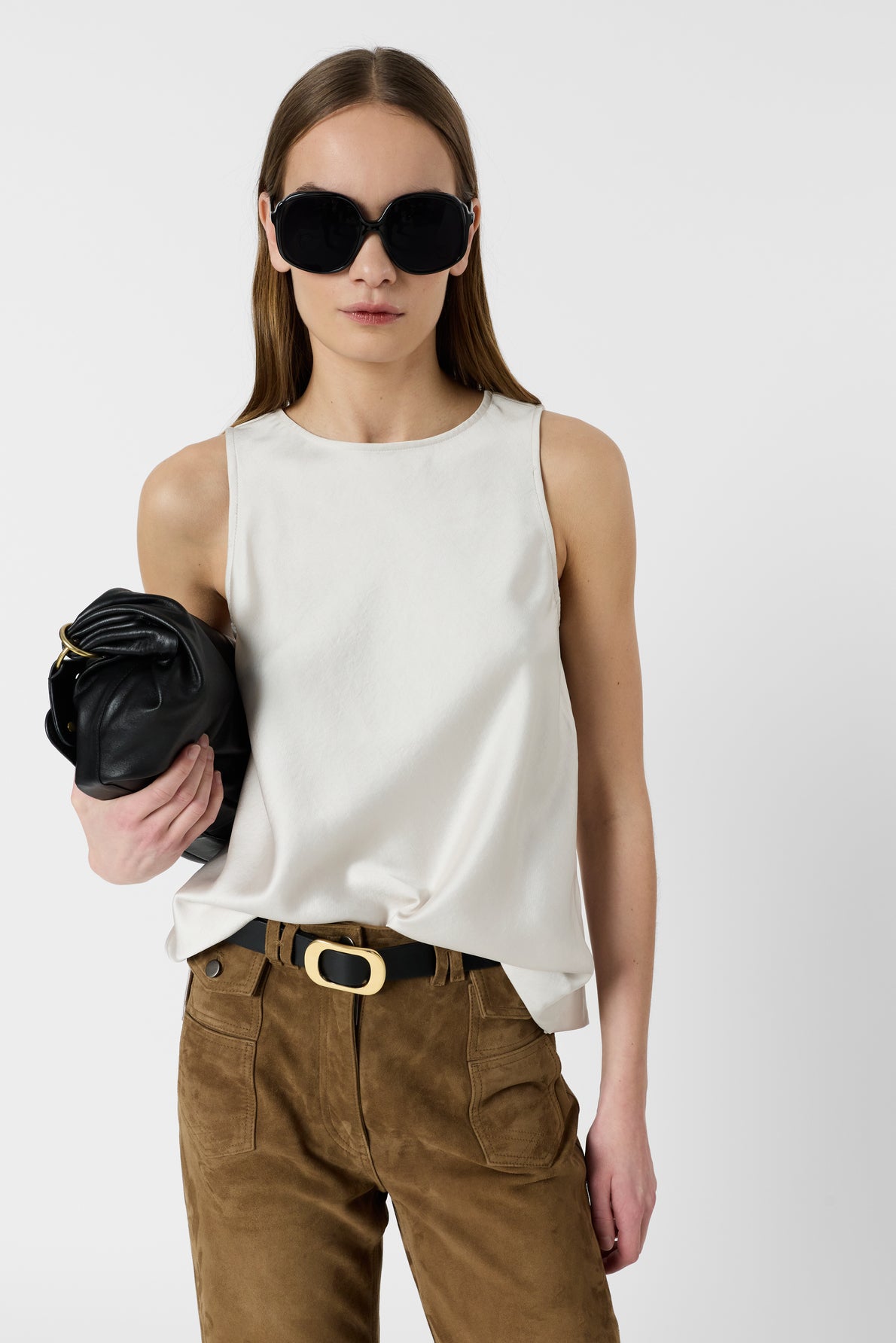 Woman wearing large black sunglasses, a sleeveless white top, and brown pants with a black belt, holding a black clutch against a plain white background.