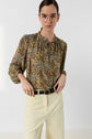 Person wearing glasses, a blouse Carinne à imprimé floral with long sleeves, and light-colored pants with a black belt, standing against a plain white background, looking directly at the camera. This édition limitée piece adds elegance to the overall look.