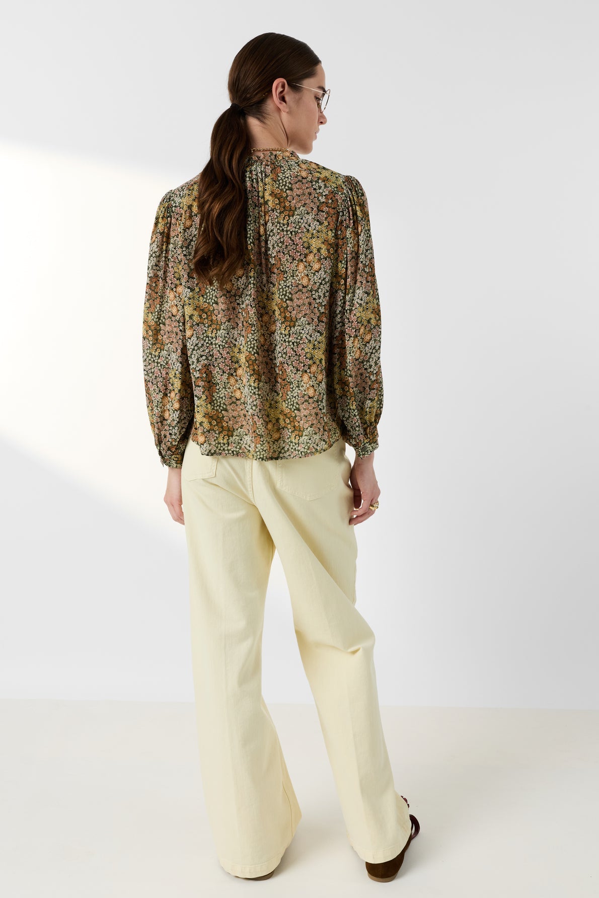 A person with long brown hair in a ponytail wears glasses, an imprimé floral blouse Carinne with long sleeves, and cream wide-leg pants, standing facing away against a plain white background.