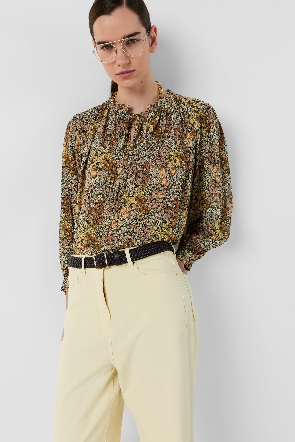 Woman wearing glasses, a blouse Carinne en édition limitée with an élégant imprimé floral and ruffled collar, paired with high-waisted cream-colored pants and a black belt, standing against a plain light gray background.