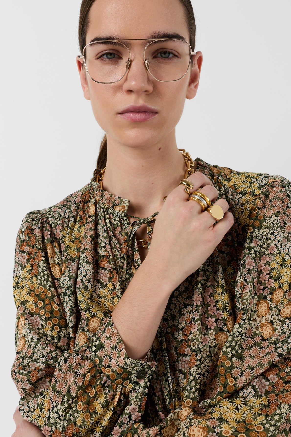 A person with straight brown hair, wearing glasses and a blouse Carinne à édition limitée with an imprimé floral pattern, poses with their hand near their chin, displaying several gold rings and a chunky chain necklace against a plain white background.