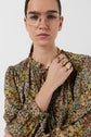 A person with straight brown hair, wearing glasses and a blouse Carinne à édition limitée with an imprimé floral pattern, poses with their hand near their chin, displaying several gold rings and a chunky chain necklace against a plain white background.