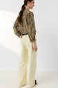 A woman with long brown hair in a ponytail wears the blouse Carinne, an imprimé floral design and part of our édition limitée collection, paired with high-waisted, wide-leg cream pants. She stands with her back to the camera against a plain white background.