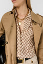 Woman wearing a tan trench coat over a patterned blouse, gold chain necklaces, and light pants with a black belt, holding a black bag.