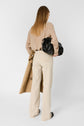 A woman with straight, light brown hair, wearing a patterned beige top and cream pants, holds a tan coat and black bag while standing against a white background, facing away from the camera.