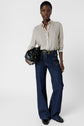 A person stands facing forward, wearing a chemise fluide élégante—a patterned button-up shirt—paired with high-waisted dark blue jeans and black shoes, holding a black handbag in one hand against a plain white background.