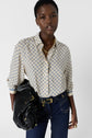 A woman wearing a chemise monogramme femme, dark high-waisted jeans, and gold jewelry holds a black handbag over her shoulder, standing against a plain white background.