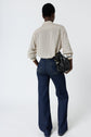 Person standing with back to camera, wearing a chemise fluide élégante patterned long-sleeve shirt, dark blue flared jeans, and holding a black handbag in their left hand against a plain white background.