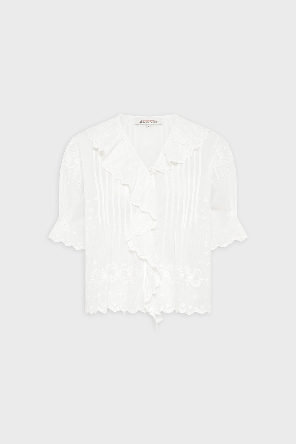 White short-sleeved blouse with ruffled collar and front, delicate floral embroidery, and scalloped edges on sleeves and hem. Set against a plain light gray background.