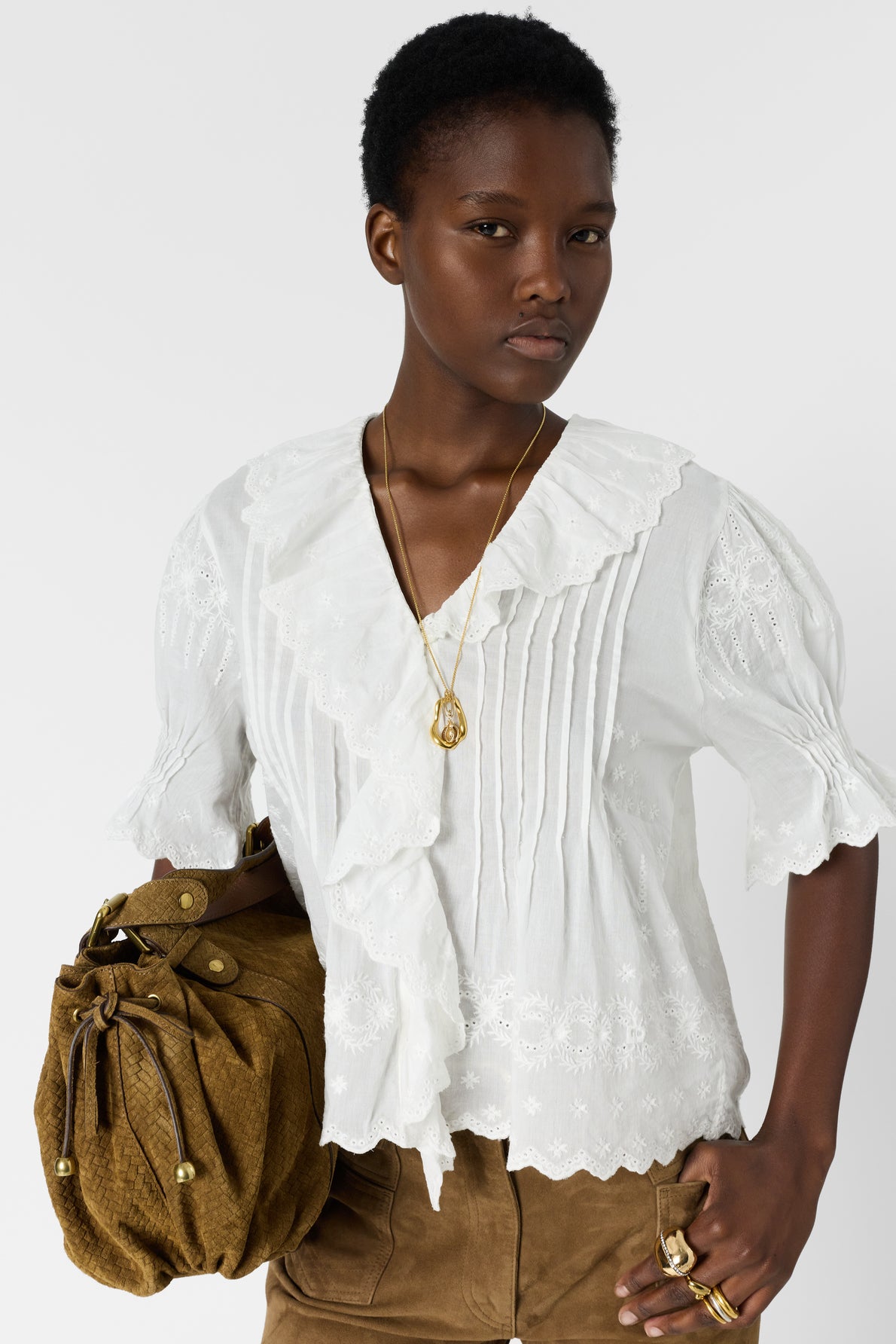 A person wearing a white ruffled blouse, brown skirt, gold necklace, and multiple rings holds a brown handbag against a plain white background.