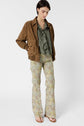Woman wearing a brown suede jacket, green ruffled blouse, floral flare pants, and sandals stands against a plain white background, looking down with hands in jacket pockets.