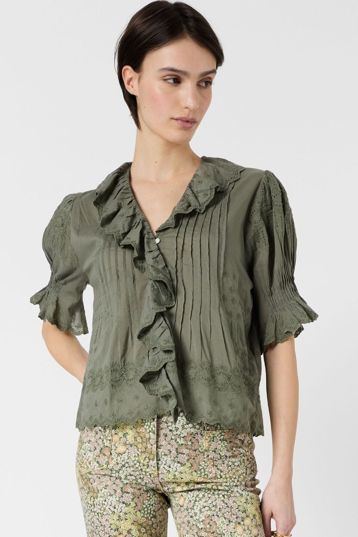 Woman wearing an olive-green, ruffled blouse with short sleeves and floral-patterned pants, standing against a plain white background.